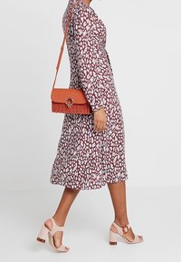 Langemouwige midi-jurk met een bruin en wit patroon, gecombineerd met een gestructureerde oranje schoudertas en lichtroze block-heeled sandalen.