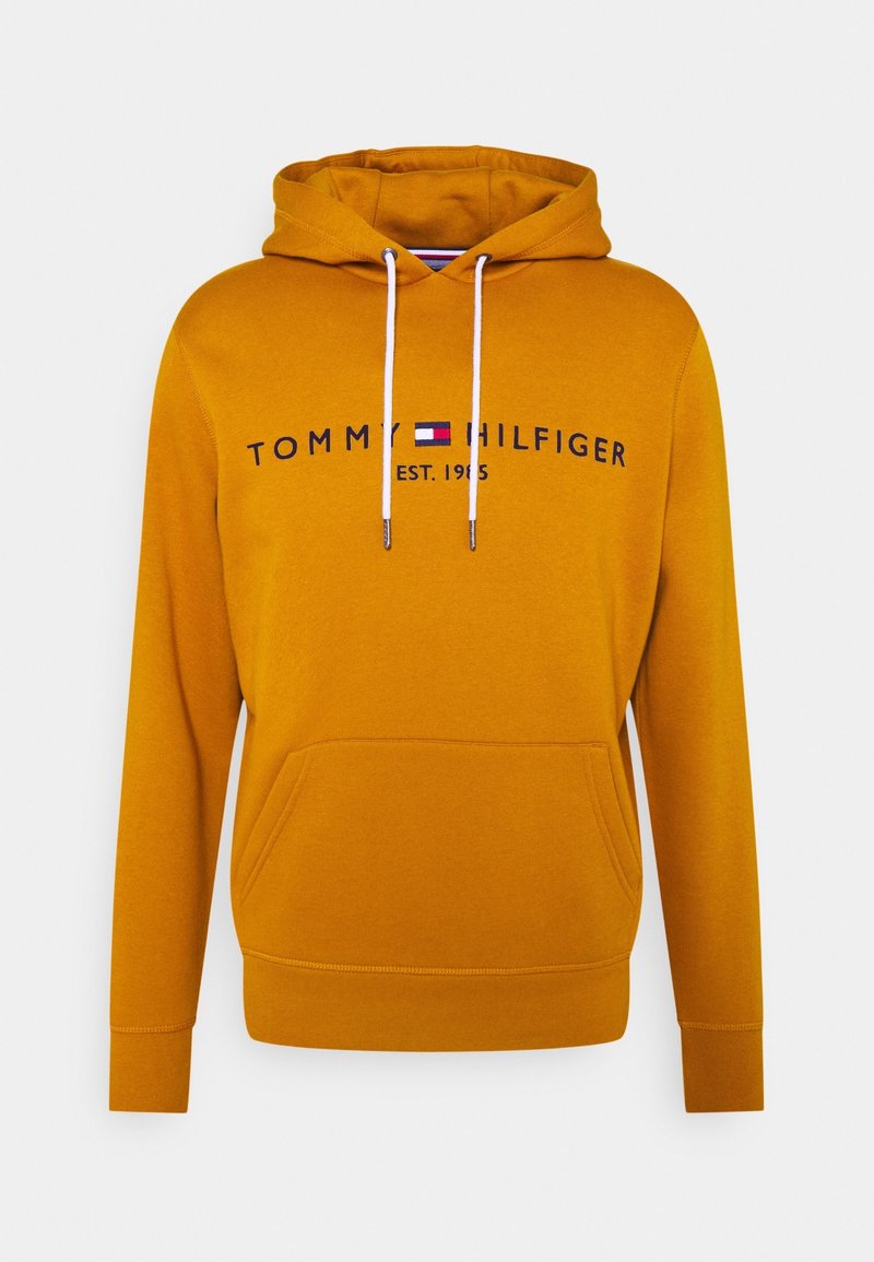 Sudadera color mostaza con bolsillo frontal, cordones y logo bordado "TOMMY HILFIGER EST. 1985" en azul marino. Tejido suave y texturizado.
