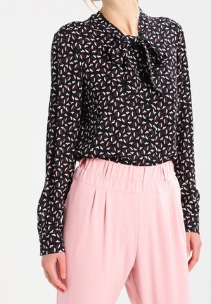 Femme portant un chemisier noir à manches longues avec un motif floral rose et blanc et un nœud au cou, associé à un pantalon rose taille haute.