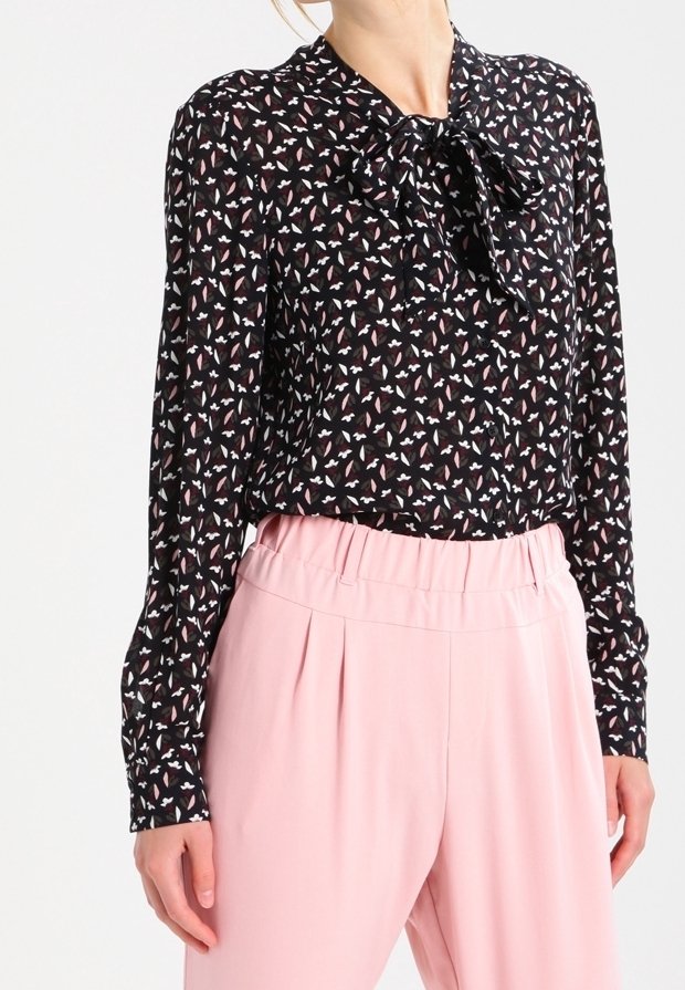 Femme portant un chemisier noir à manches longues avec un motif floral rose et blanc et un nœud au cou, associé à un pantalon rose taille haute.