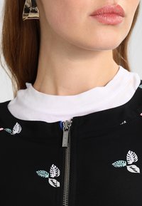 Svart jacka med blommig broderi med gröna och rosa blad, dragkedja, lager på en vit t-shirt med rund halsringning.