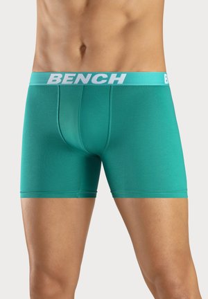 Man wearing teal boxer briefs with "BENCH" white logo on the waistband, standing against a plain light background.