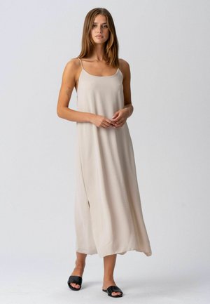 Egyenes barna hajú nő laza, ujjatlan, bézs maxi ruhát és fekete nyitott orrú papucsot visel, egyszerű háttér előtt áll.