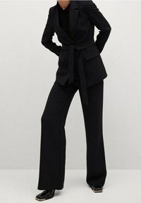 Traje negro a medida que presenta una chaqueta entallada con cinturón, pantalones rectos y una tela texturizada. Los detalles incluyen bolsillos laterales y zapatos elegantes.