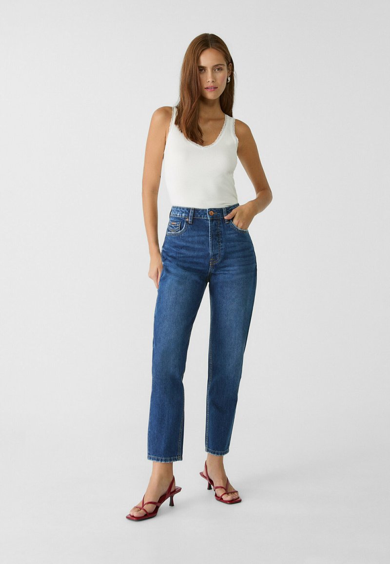 Hochgeschnittene blaue Denim-Jeans mit geradem Bein, Knopfverschluss und Vordertaschen, kombiniert mit einem weißen Tanktop und roten Riemchen-Heels.