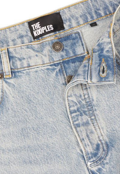 Jean en denim délavé bleu clair avec une étiquette noire "THE KOOPLES" et des boutons métalliques embossés du texte "THE KOOPLES", braguette boutonnée partiellement ouverte.