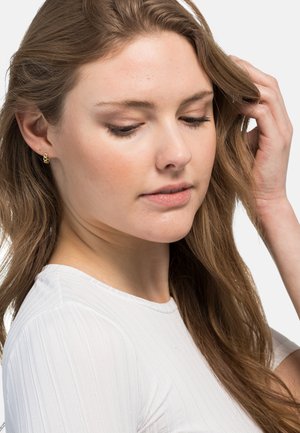 Goldene Creolen, die von einem Modell getragen werden, mit einem schlichten Design. Das Modell hat lange, wellige, braune Haare und trägt ein weißes, geripptes Oberteil.