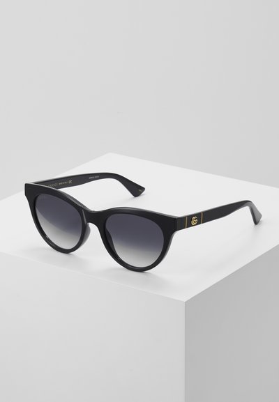 Gafas de sol negras con montura redonda, lentes oscuros en degradé y detalle del logotipo GG dorado en la patilla. Fabricadas con material plástico brillante.