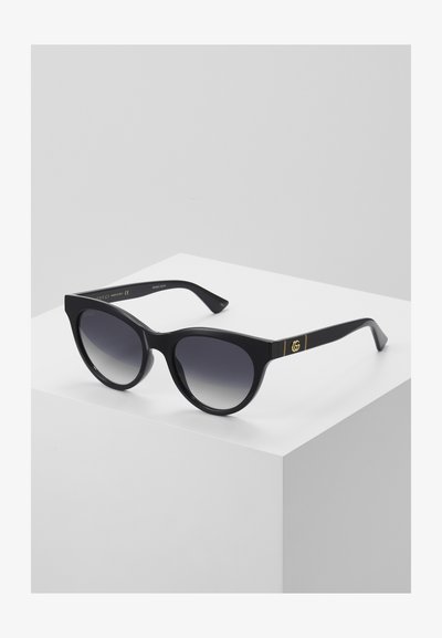 Gafas de sol negras con montura redonda, lentes oscuros en degradé y detalle del logotipo GG dorado en la patilla. Fabricadas con material plástico brillante.