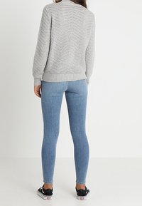 Grauer Strickpullover mit Chevron-Struktur, kombiniert mit hellblauen Skinny-Jeans. Dezente gerippte Bündchen und Saum, getragen mit schwarzen Sneakern.