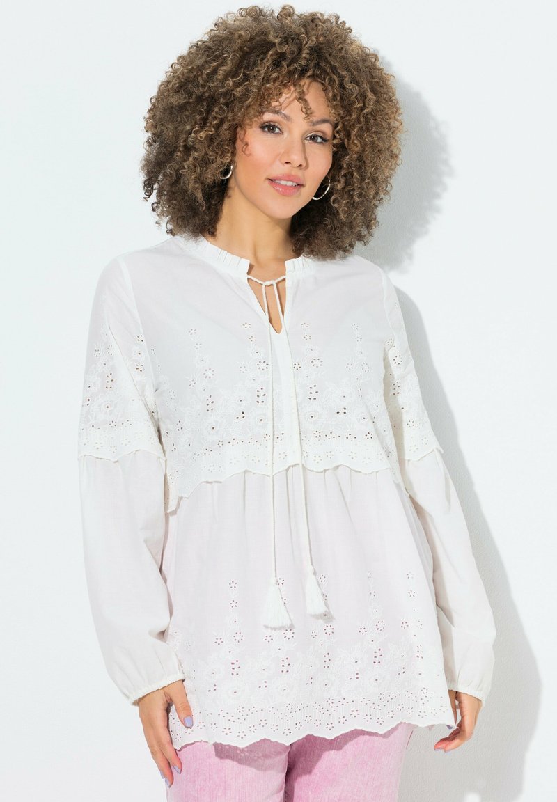 Vrouw met krullend haar, gekleed in een witte blouse met lange mouwen en kantdetails en een roze broek, staand tegen een witte achtergrond.