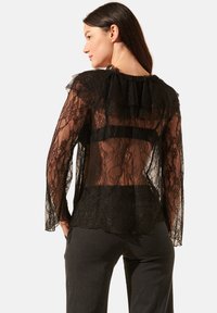 Blouse en dentelle noire avec des manches longues, un col froncé, un tissu transparent et un design superposé avec un sous-vêtement noir uni.