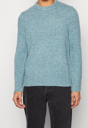 Mann trägt einen hellblauen, gesprenkelten Strickpullover mit langen Ärmeln und gerippten Bündchen, kombiniert mit schwarzen Jeans.
