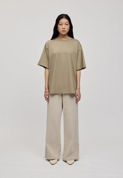 Tricou oversized din bumbac bej, asortat cu pantaloni largi bej deschis și pantofi crem cu vârful ascuțit; modelul stă în fața unui fundal neutru.