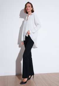 Weiße langärmlige Bluse mit Knopfverschluss und abgerundetem Saum, kombiniert mit schwarzen, taillierten Hosen und spitzen schwarzen Absätzen auf einem hellen Holzboden.
