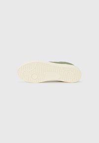 Sola de sapatilha em camurça verde com uma sola de borracha branca texturizada, apresentando um detalhe de logotipo circular e um padrão de piso para aderência.