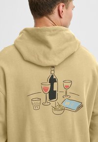 Mann iført en beige hettegenser med tegning av en vinflaske, to vinglass, et glass med vann, en bolle med oliven og en bok på baksiden.
