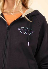 Haut à capuche noir avec fermeture éclair à l'avant, doublure de capuche crème et broderie colorée "O.X.B.C.W". Présente des accents violets et aquatiques sur le design.