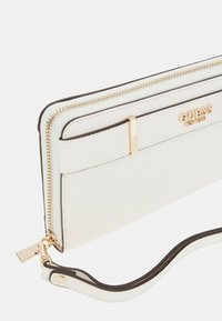 Portafoglio bianco in ecopelle con accessori in oro, chiusura con zip e tracolla. Presenta un accento marrone lungo i bordi e il logo del marchio.