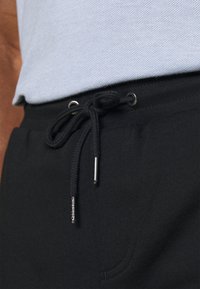 Sorte joggingbukser med snor i taljen, metaldetaljer og en tekstureret stof. Sidelomme er synlig.