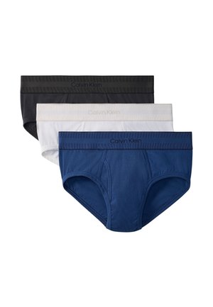 Jeune homme debout pieds nus sur un sol en béton, portant un slip noir Calvin Klein sur un fond gris clair uni.