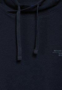 Námořnická modrá mikina s kapucí vyrobená z texturované tkaniny, s tkaničkami a malou tištěnou etiketou s bílým textem na boku.