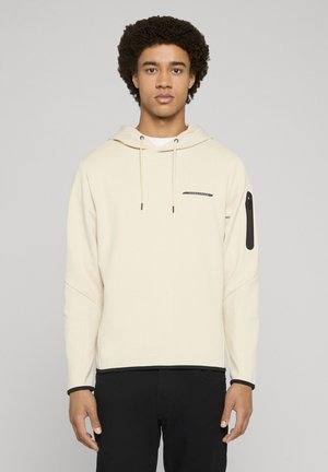 Jonge man met krullend haar, draagt een crèmekleurige hoodie met een zak aan de voorkant en zwarte broek, tegen een effen lichte achtergrond.