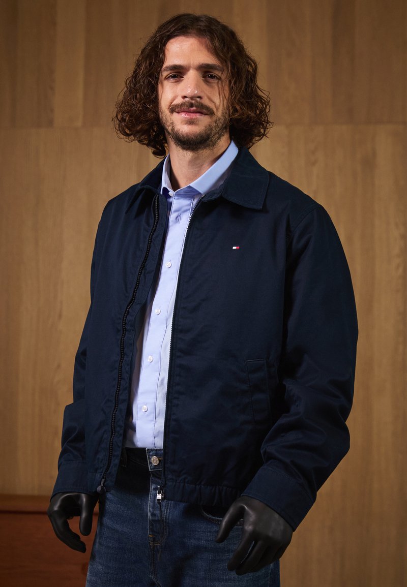 Marine bomberjakke med glidelåsfront, som har krage, sidelommer og ribbestrikket nederkant. Båret over en lyseblå skjorte og mørke jeans.