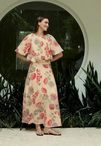 Blumiges zweiteiliges Outfit mit kurzen Ärmeln, das rote und blaue Blumen auf einem hellbeigen Stoff zeigt. Langer Rock und flache goldene Sandalen.