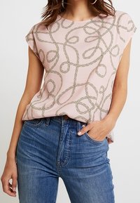 Light pink short-sleeve top with a chain pattern in gold. Hemline is slightly curved, and it’s paired with high-waisted blue jeans.