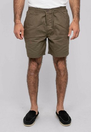 Homme portant un short vert olive à cordon, chemise blanche, chaussures noires sans lacets, debout sur un fond gris uni.
