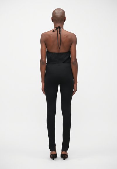 Zwarte halter jumpsuit met een aangesloten lijfje, strakke leggings en een strikdetail aan de achterkant. Soepele textuur, minimalistisch ontwerp en enkels hoge splits.