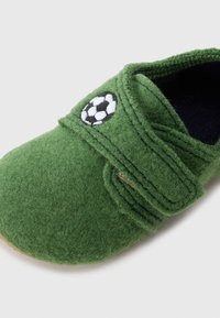 Zeleni filcasti copat z Velcro trakom, s črno-belo vezenino nogometne žoge. Mehka tekstura in zaobljena oblika.
