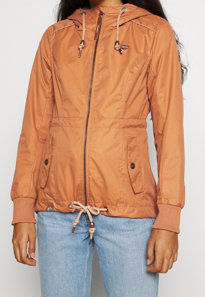 Veste marron à capuche avec fermeture éclair, taille ajustable par cordon de serrage et poignets élastiques. Elle comprend deux poches avant et un détail de surpiqûres discret.