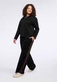 Czarna bluza z półzamkiem i sznurkami, zestawiona z czarnymi spodniami z brązowymi bocznymi paskami. Model ma na sobie jasne trampki.