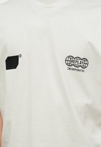 Λευκό μπλουζάκι με μικρά μαύρα λογότυπα: ένα σταθερό γεωμετρικό σχήμα με καταχωρημένο εμπορικό σήμα στο αριστερό στήθος και "REPLAY INCORPORATED" με εικονίδια γης στο δεξί στήθος.