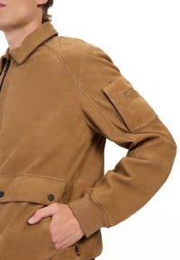 Brun læderjakke med lynlåslukning, ribbede manchetter og en frontlomme med klap. Har en skulderpatch og en glat tekstur.