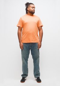 Άνδρας με ντραντλοκς που φοράει πορτοκαλί μπλουζάκι, μπλε τζιν και μαύρα αθλητικά παπούτσια, στέκεται μπροστά σε απλό λευκό φόντο, κοιτάζοντας πλάγια.