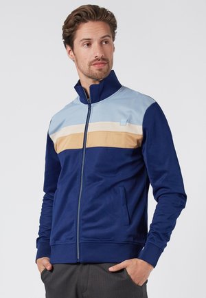 Marine blauwe zip-up jacket met lichtblauwe, beige en crèmekleurige horizontale strepen op de borst. Voorzien van zijzakken en een hoge kraag.