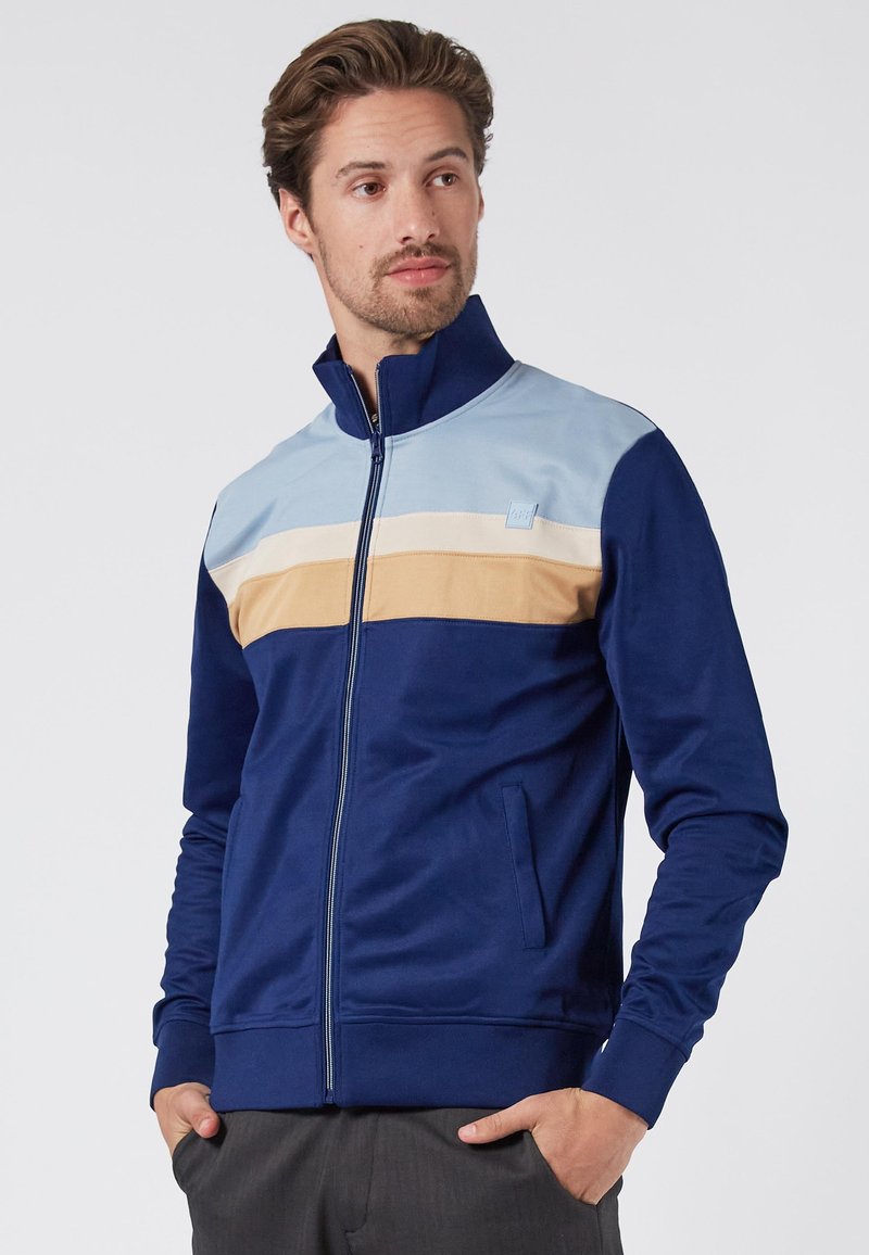 Navy zip-up jacket with light blue, beige, and cream horizontal stripes across the chest. Features side pockets and a high collar.