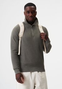 Dunkelgrauer Reißverschluss-Pullover mit Stehkragen, gerippten Bündchen und Saum. Das Modell trägt einen beigen Rucksack über beiden Schultern.