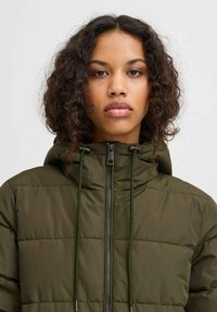 Veste matelassée vert olive avec fermeture éclair à l'avant, capuche avec cordon de serrage et fermeture contrastante. Le tissu présente une texture lisse et brillante.