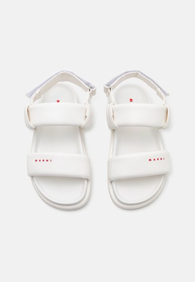 Sandalias blancas con dos correas anchas y un acabado suave y texturizado. Presenta un logo rojo en la suela, hebilla ajustable en el tobillo y forma redondeada.