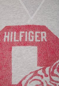 Šedá bavlněná mikina s výraznou červenou grafikou obsahující slovo "HILFIGER" a čtvercový designový prvek. Švy kolem výstřihu jsou viditelné.