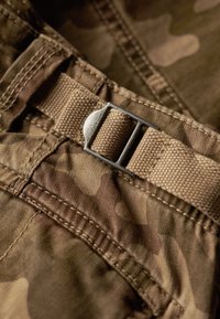 Bruin camouflage stof met een gestructureerd oppervlak, met een beige riem en metalen gesp. Close-up van de stitching en details.
