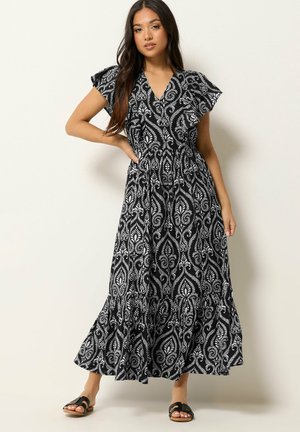 Kobieta w czarno-białej wzorzystej sukience maxi z falującymi rękawami, stojąca na jednolitym jasnym tle, w czarnych klapkach.