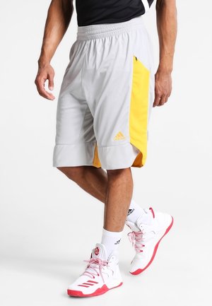 Mężczyzna ubrany w białe i żółte spodenki do koszykówki Adidas, białe skarpetki Adidas oraz białe i czerwone buty do koszykówki, stojący ze skrzyżowanymi nogami.