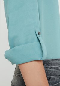 Manche longue bleu clair retroussée avec détail de bouton-pression noir au-dessus du coude, portée avec un jean denim gris foncé.