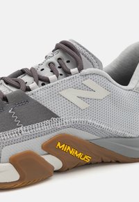 Lys grå sportsko med en struktureret mesh-overdel, mørkegrå accenter, brun sål og "MINIMUS" logoen tydeligt vist.