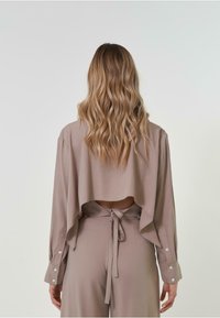 Haut de couleur brun clair, ample, à manches longues avec un dos ouvert, des détails à nouer et des poignets boutonnés. Tissu doux avec une texture lisse.